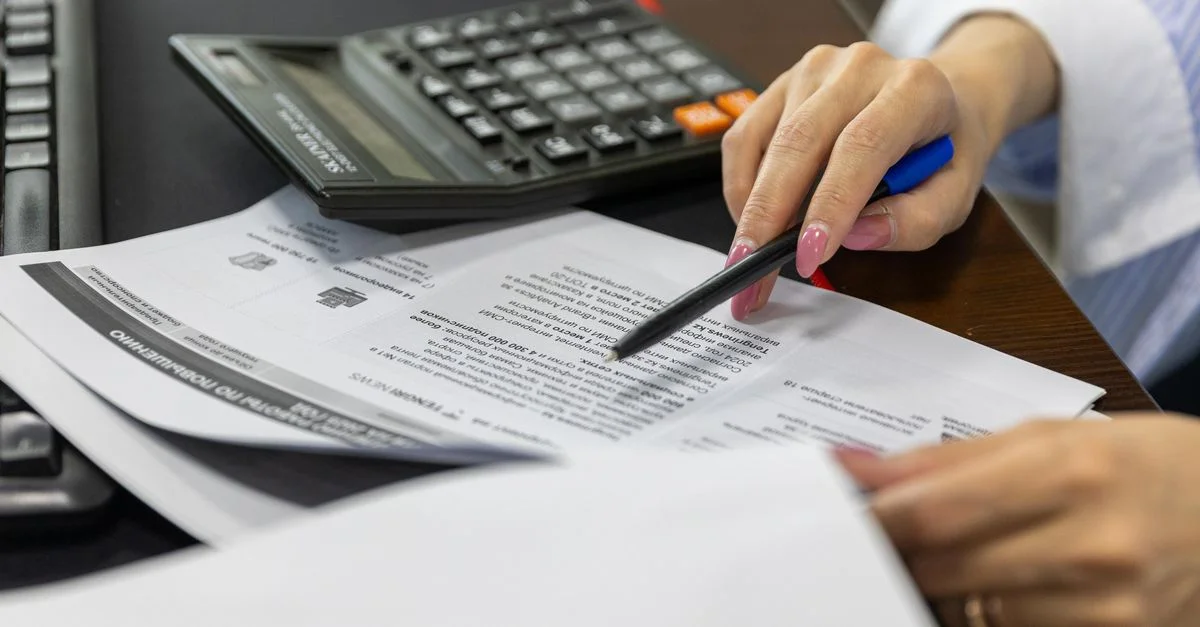 Primer plano de la mano de una mujer revisando documentos financieros con una calculadora.
