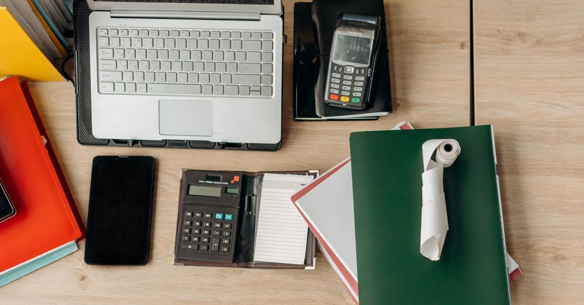 Un escritorio de oficina organizado con un portátil, calculadora, terminal de pago y documentos.