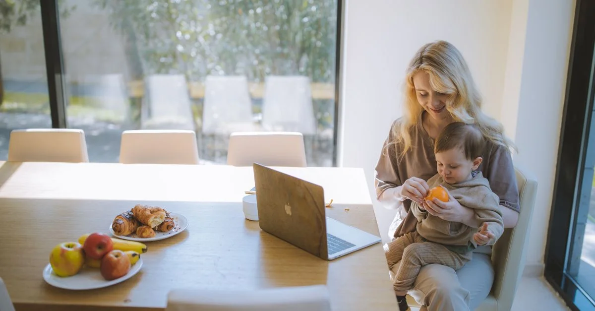 Madre rubia sosteniendo al bebé con una laptop en la mesa del comedor, disfrutando del desayuno en interiores.