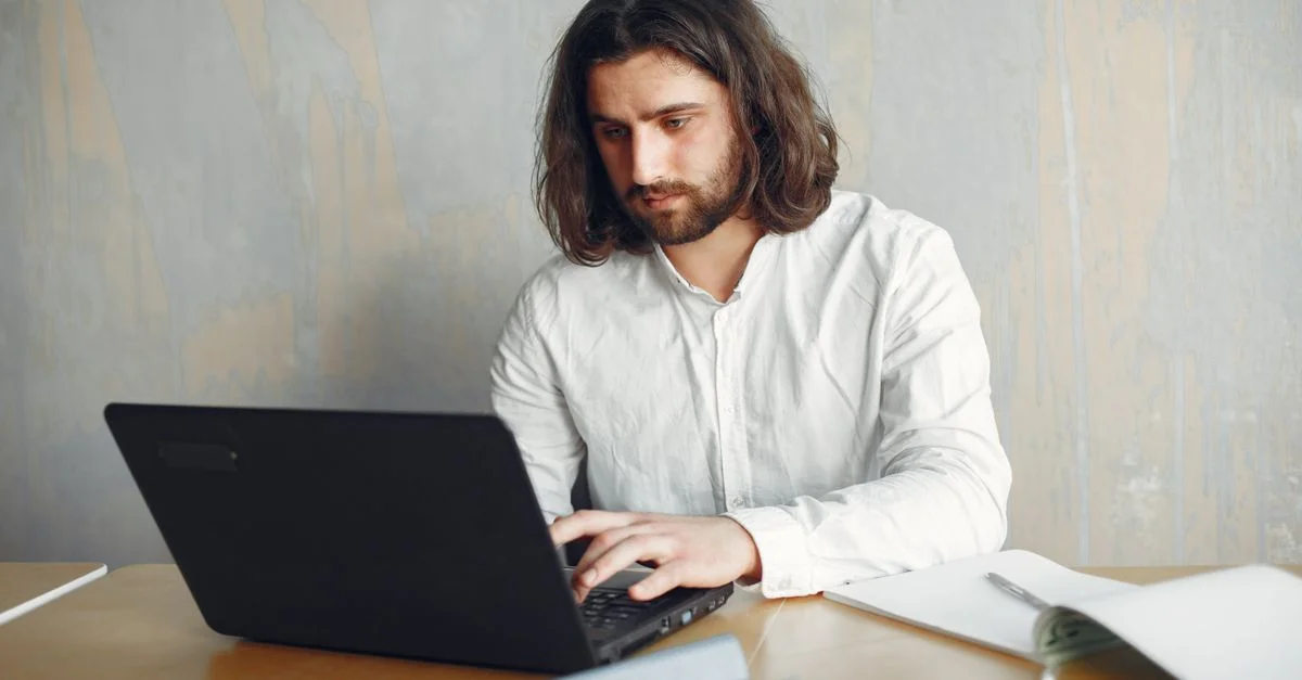 Un hombre con barba trabajando de forma remota en su laptop en un escritorio con documentos, mostrando un entorno de trabajo moderno.