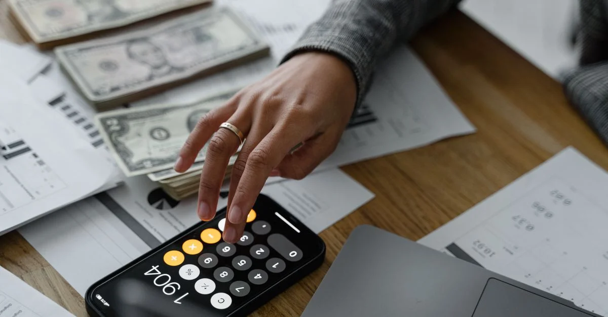 Gros plan sur une personne utilisant une calculatrice sur smartphone parmi de l'argent et des documents financiers sur une table en bois.