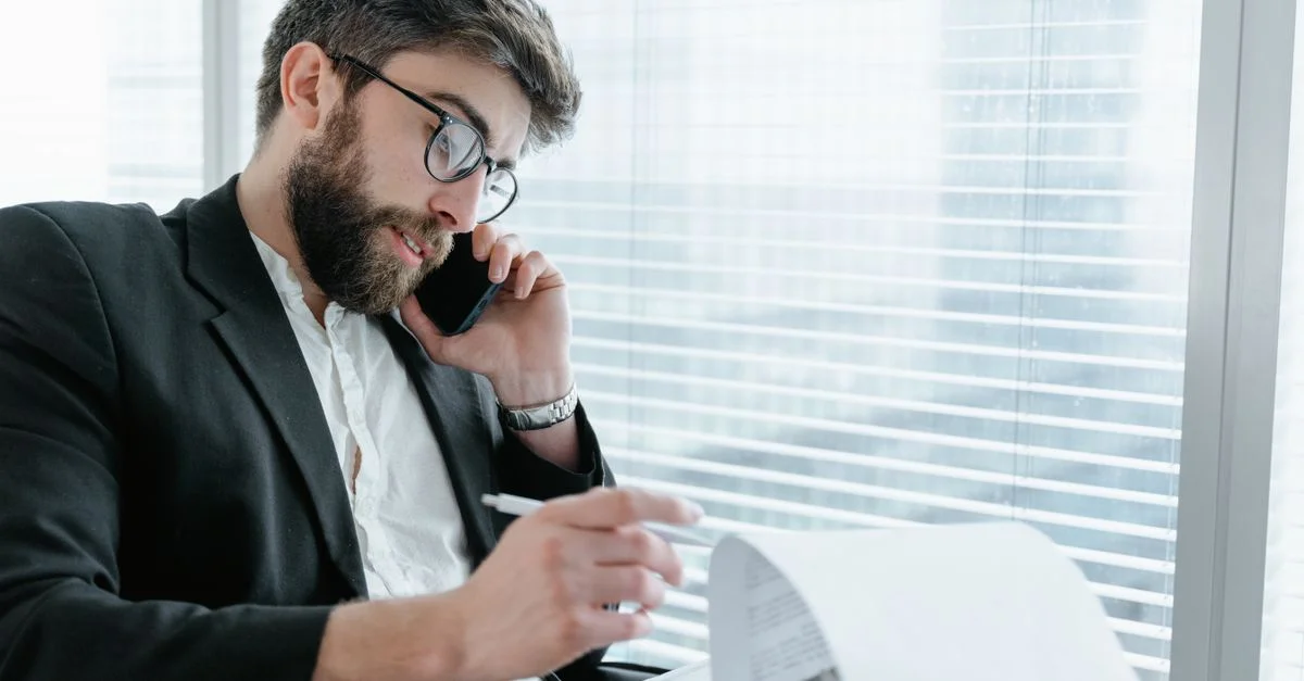 Hombre de negocios con gafas haciendo una llamada telefónica mientras revisa documentos en un entorno de oficina moderno.