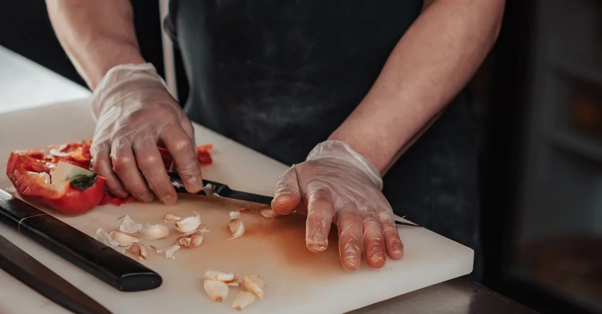 Un chef portant des gants tranche de l'ail et du poivron rouge sur une planche à découper, mettant en avant la précision et l'hygiène.