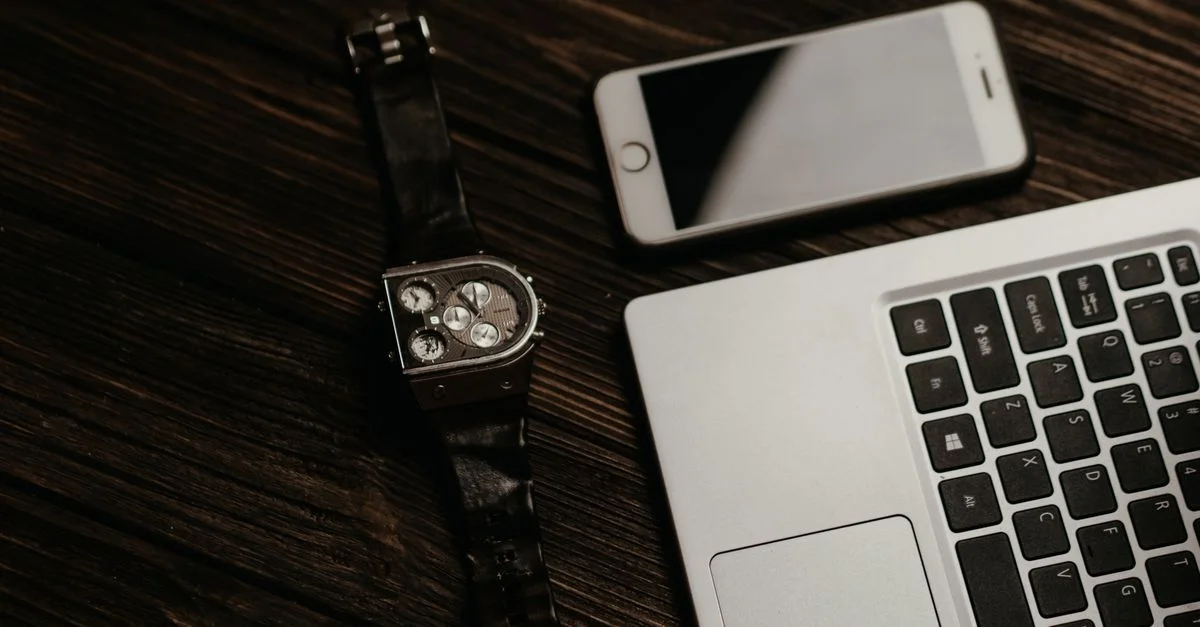 Primer plano de un laptop, smartphone y reloj de pulsera sobre una mesa de madera, capturando una elegante configuración de oficina.
