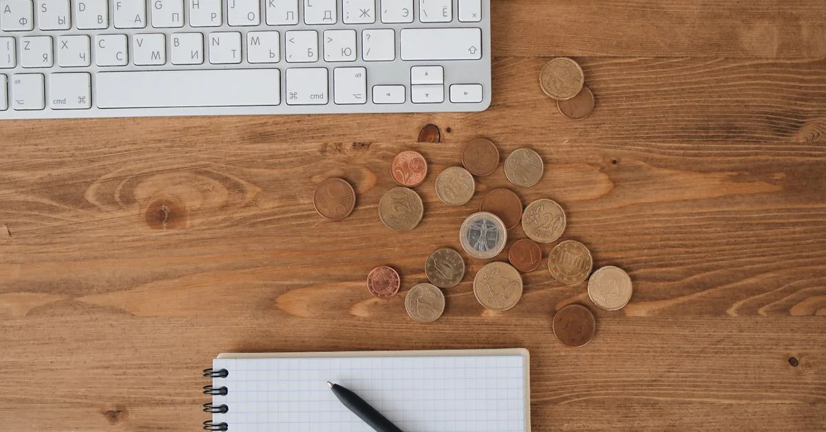 Monedas esparcidas sobre un escritorio de madera junto a un teclado y un cuaderno, capturadas desde arriba.