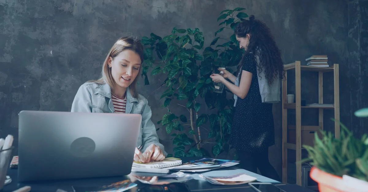 Dos mujeres trabajando en una oficina elegante con portátiles y vegetación.