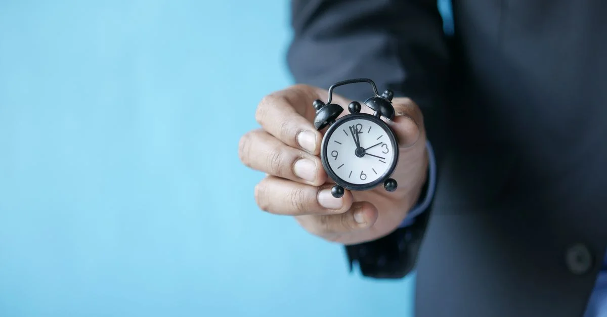 Primer plano de la mano de una persona sosteniendo un pequeño reloj despertador analógico negro sobre un fondo azul.
