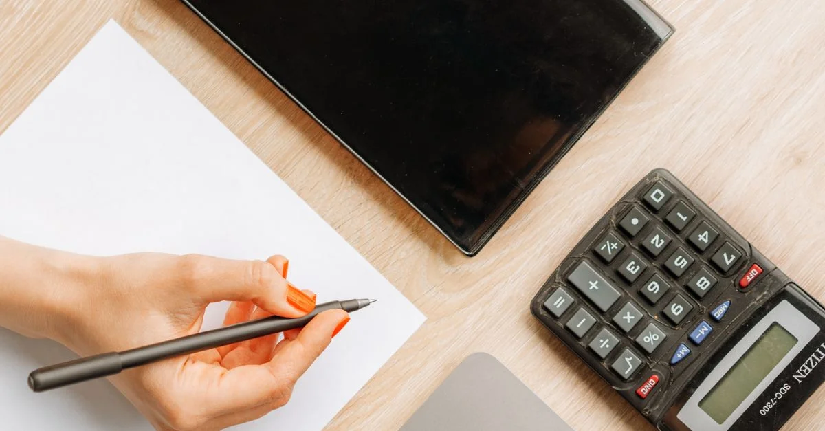 Vista superior de un escritorio minimalista con una mano escribiendo, calculadora y dispositivo electrónico.