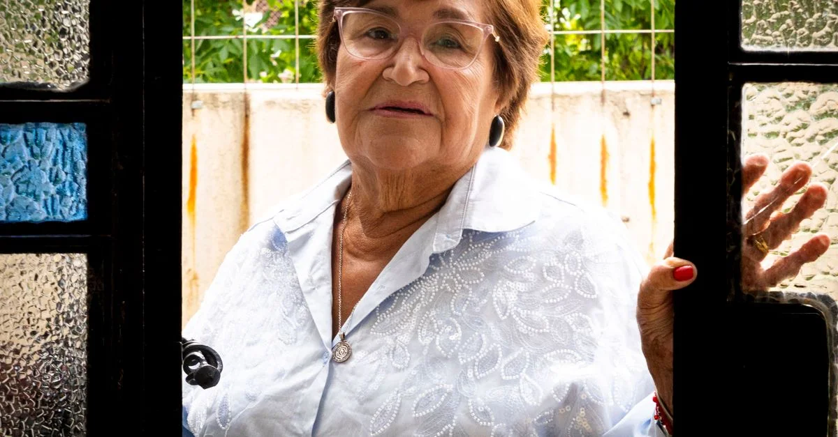 Una mujer mayor con gafas está de pie en el umbral de una puerta, mostrando una expresión serena.