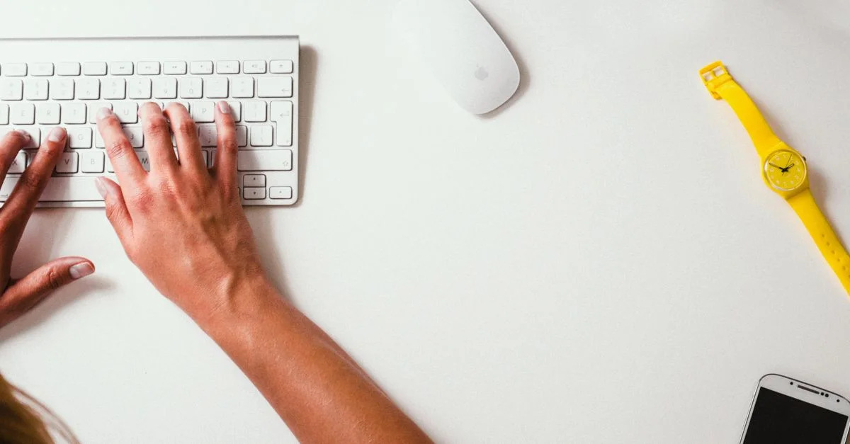Primer plano de manos escribiendo en un teclado con un reloj amarillo en la mesa