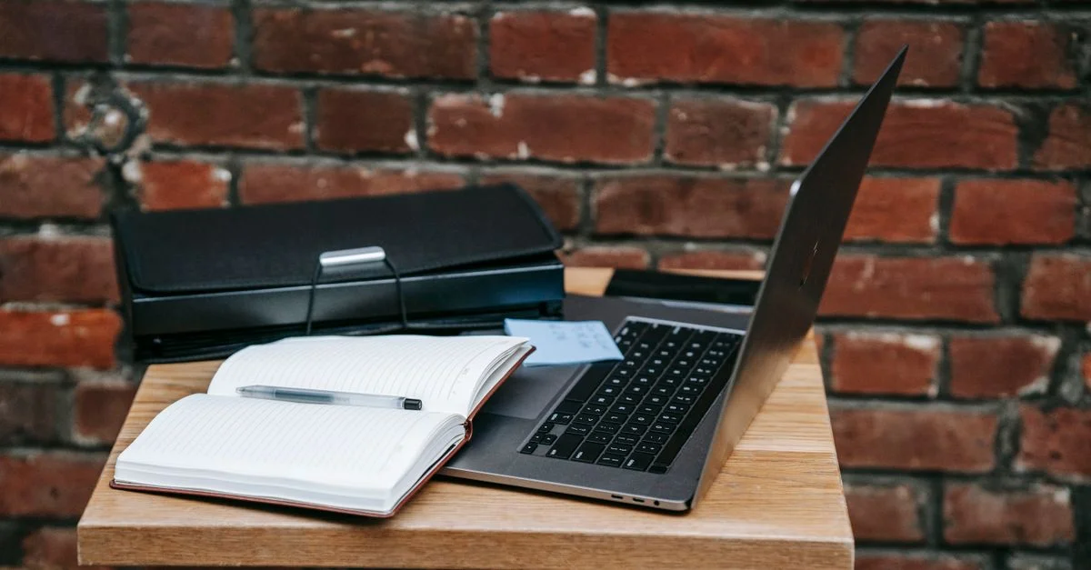 Portátil abierto y cuaderno sobre una mesa de madera junto a una pared de ladrillo, perfecto para inspiración de trabajo remoto.