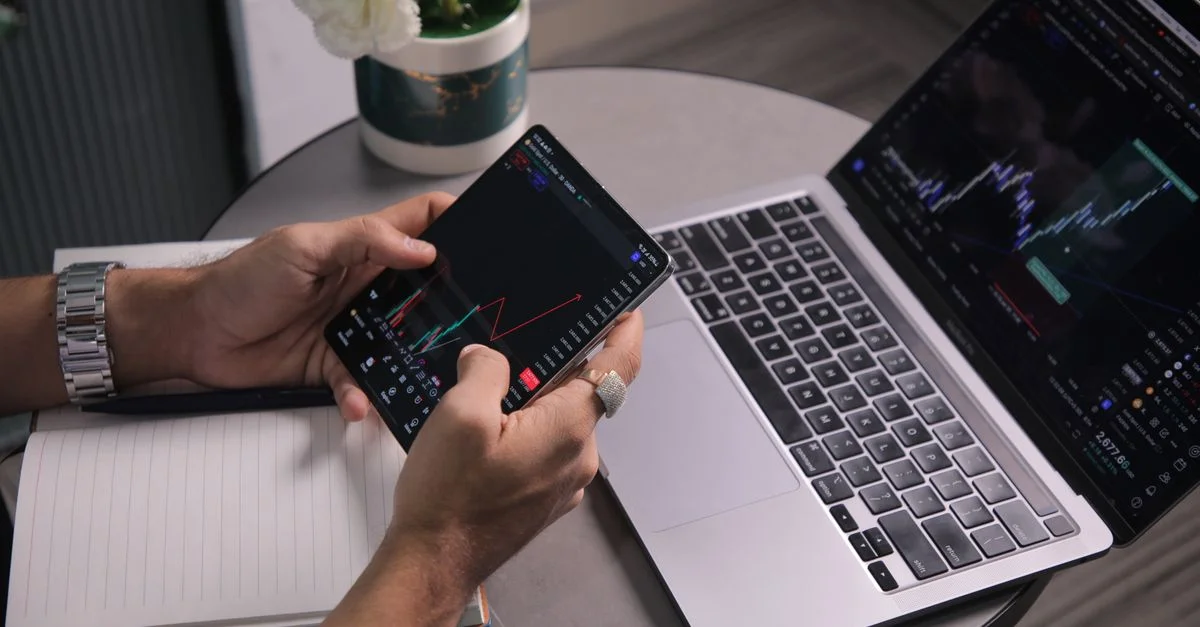 Un homme analyse les tendances boursières avec un ordinateur portable et une application de trading mobile dans un espace de travail moderne.