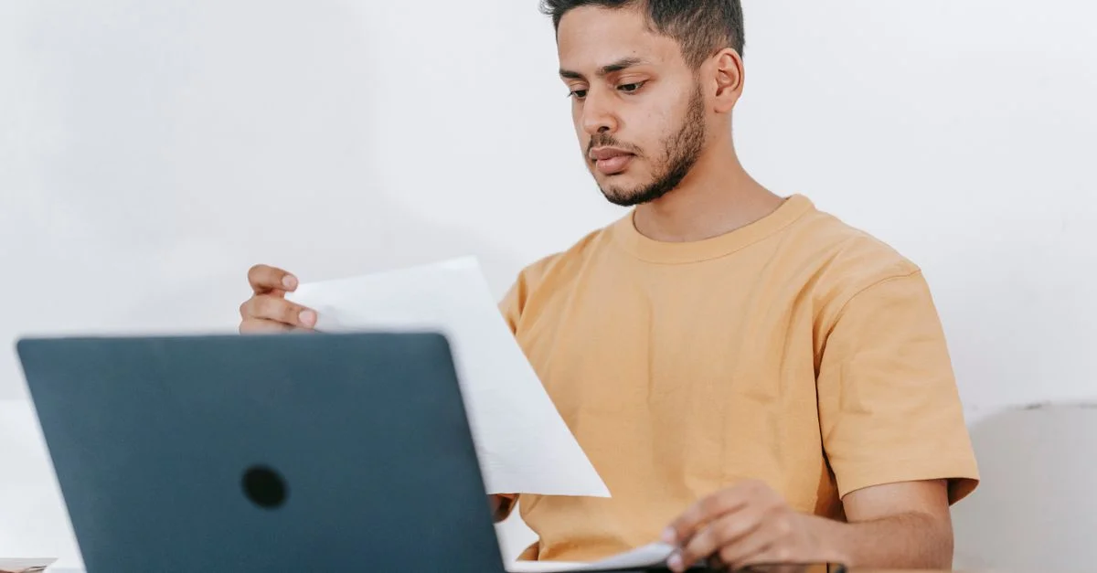 Un joven examina atentamente documentos mientras trabaja en su laptop en un entorno de oficina en casa.